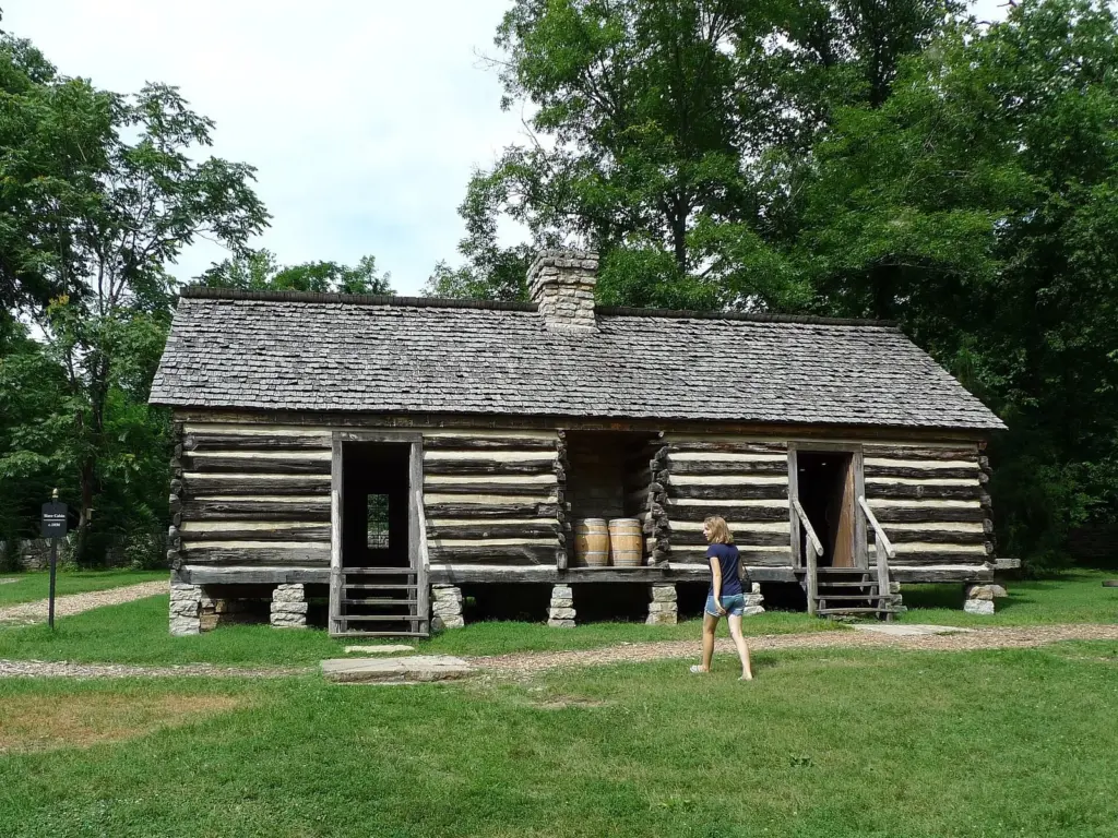 A reconstructed log cabin with two doors and a stone chimney on the lawn at Belle Meade, with a visitor walking past
