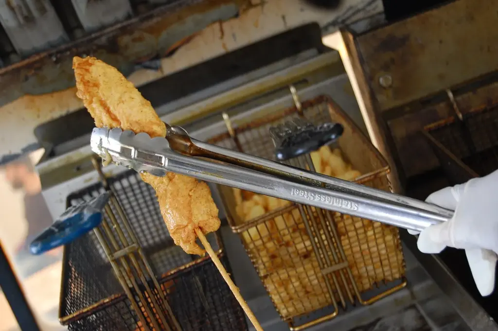 A piece of hot chicken on a stick being lifted out of the fryer with tongs at Bolton's Spicy Chicken and Fish