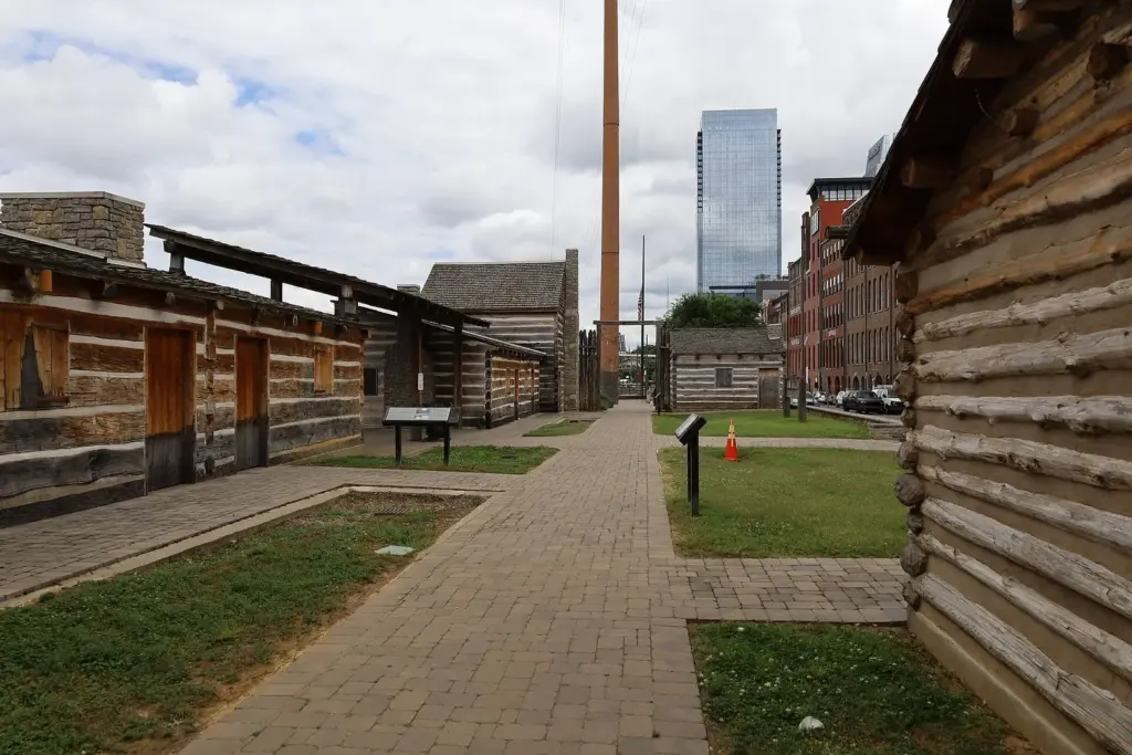 Log cabins and a paved path inside Fort Nashborough in Riverfront Park, one of the free things to do in Nashville, with downtown towers behind