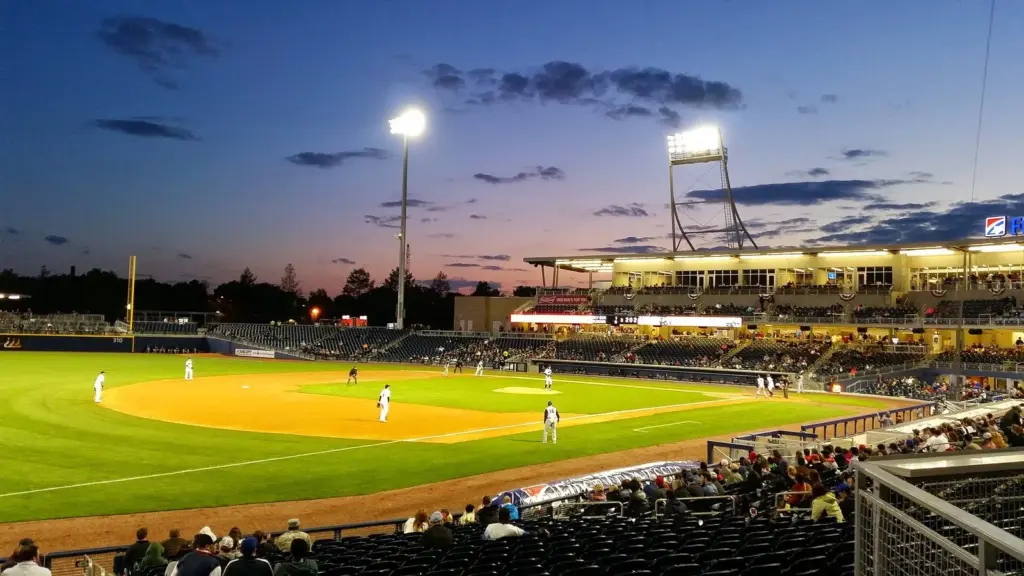 A night baseball game under lights at the Nashville Sounds ballpark at dusk, with fans filling the lower seats