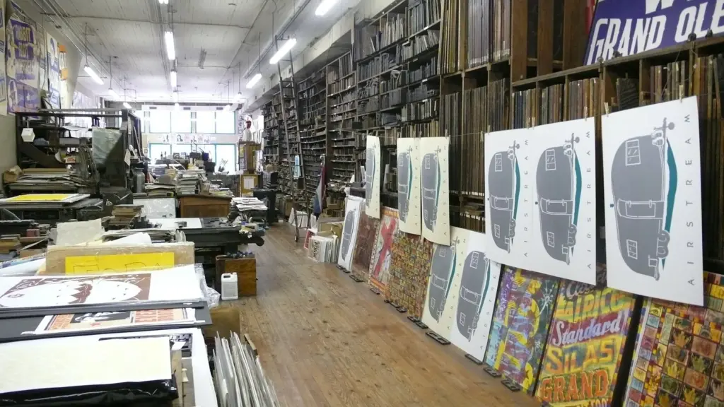 Letterpress posters drying along the walls of a print shop, with shelves of wood type behind them